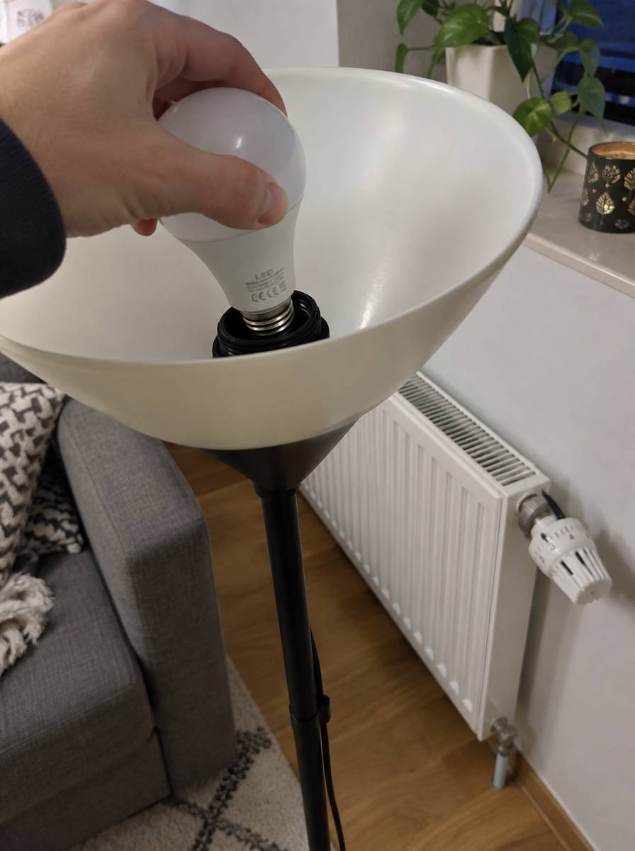 Dłoń wkręca żarówkę LED w lampę, w tle grzejnik z termostatem w mieszkaniu